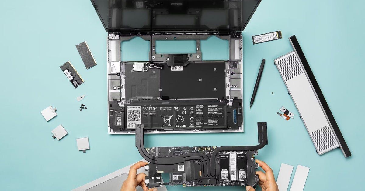 A close-up of a modular Framework Laptop 16 motherboard showing exposed RAM slots on a wooden desk with tools.