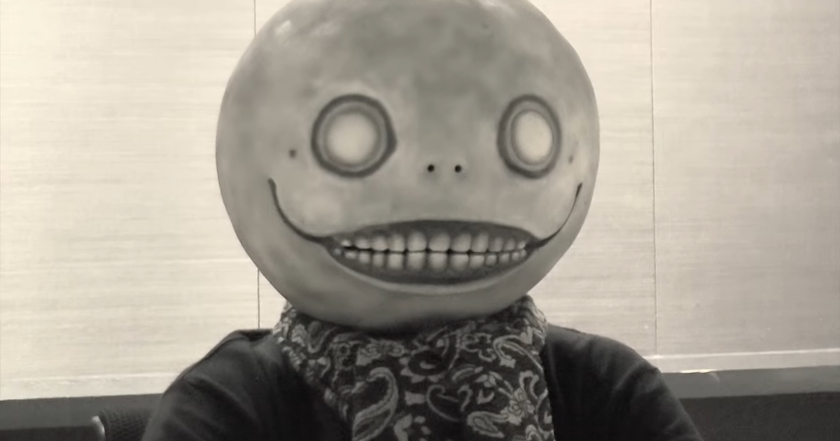 alt="Yoko Taro wearing his signature Emil mask standing next to Evangelion Unit 01 against a red apocalyptic sky"